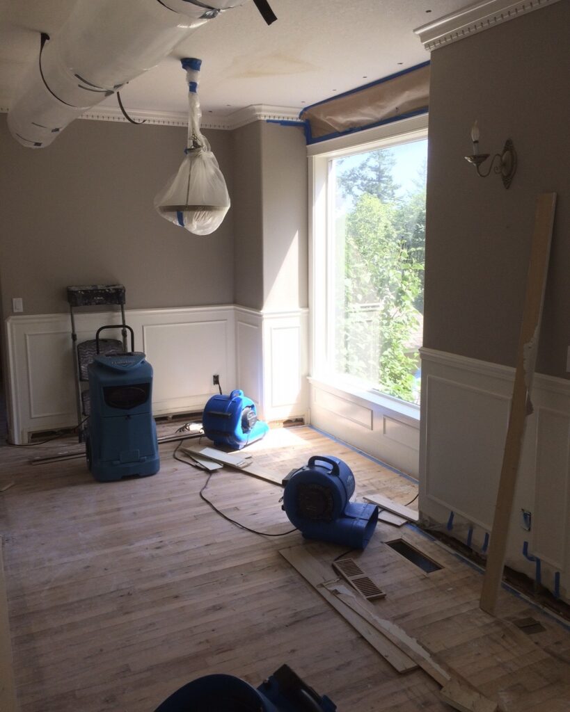 A living room with a big window and unfinished hardwood floors in Camas WA with a water damage restoration dehumidifier and centrifugal fans